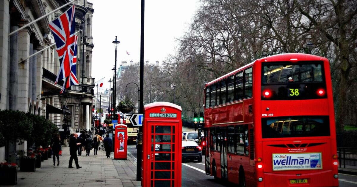 Foto de rua em Londres, com cabine, bandeiras e um ônibus de dois andares