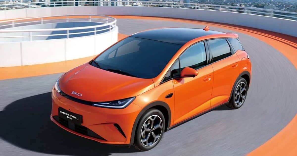 BYD Dolphin G laranja em movimento em uma pista circular, com visual moderno e teto panorâmico preto, tendo ao fundo uma paisagem urbana com prédios e arranha-céus