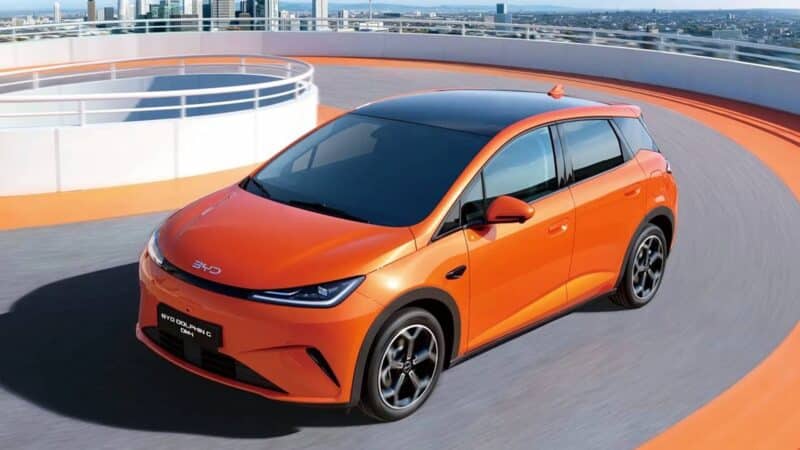BYD Dolphin G laranja em movimento em uma pista circular, com visual moderno e teto panorâmico preto, tendo ao fundo uma paisagem urbana com prédios e arranha-céus