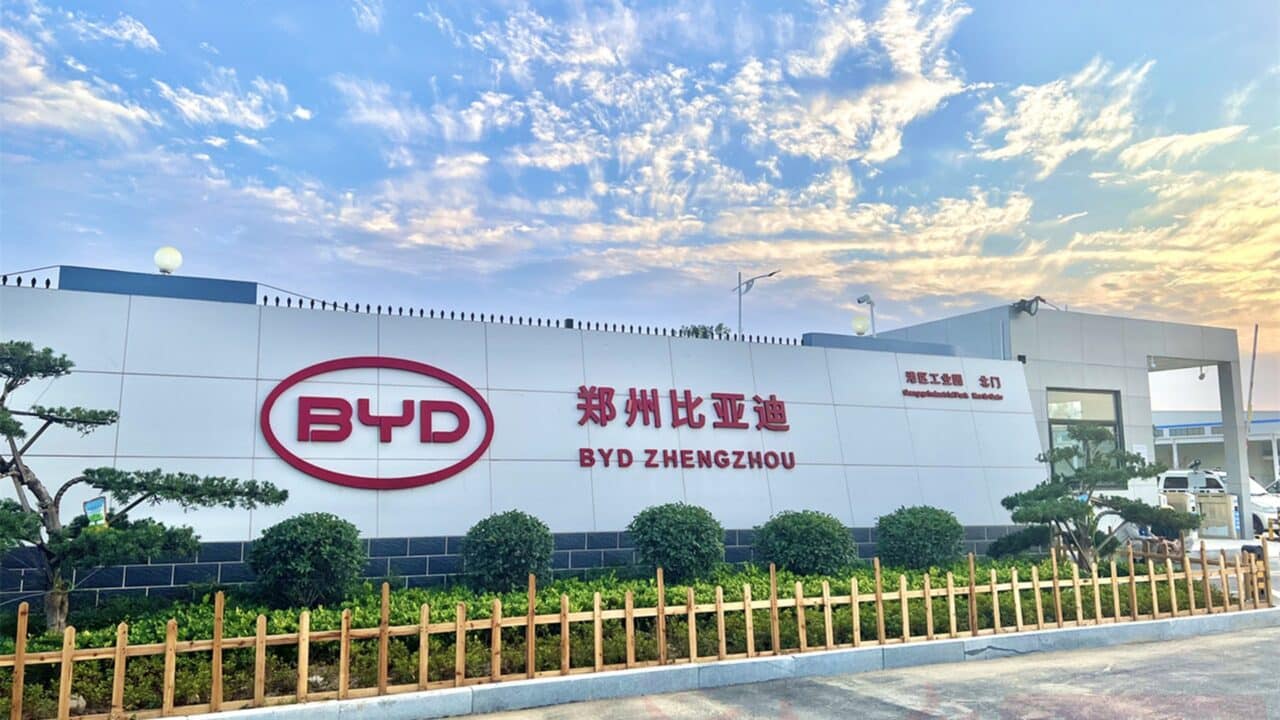 Fachada externa da unidade industrial da BYD localizada em Zhengzhou, China. O edifício é amplo, de estrutura moderna, com paredes brancas. Em destaque na parede central, o logotipo da BYD em vermelho