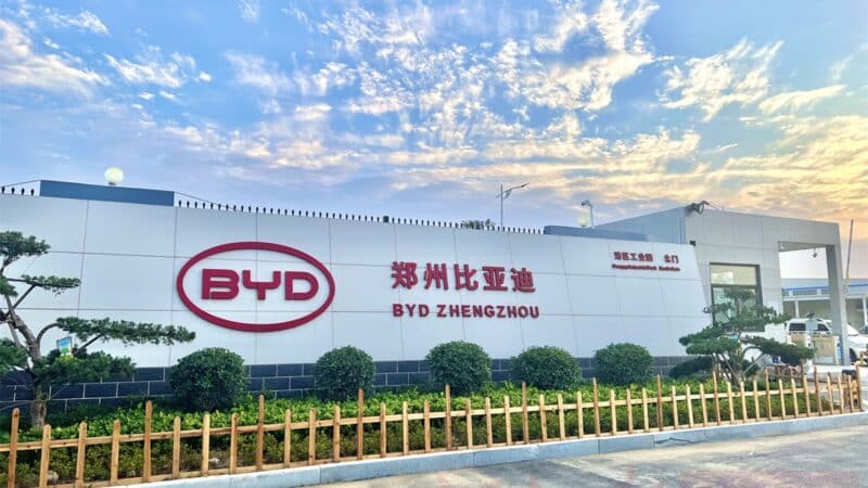 Fachada externa da unidade industrial da BYD localizada em Zhengzhou, China. O edifício é amplo, de estrutura moderna, com paredes brancas. Em destaque na parede central, o logotipo da BYD em vermelho