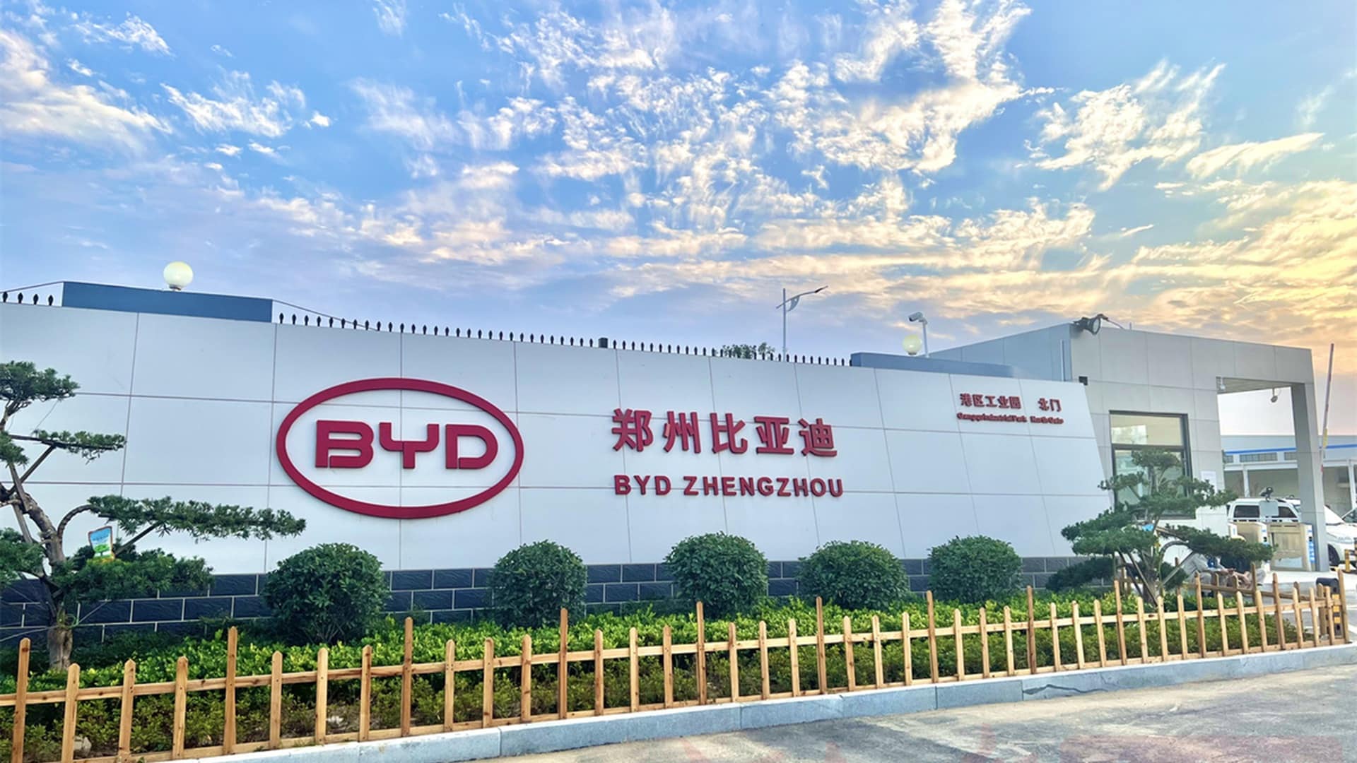 Fachada externa da unidade industrial da BYD localizada em Zhengzhou, China. O edifício é amplo, de estrutura moderna, com paredes brancas. Em destaque na parede central, o logotipo da BYD em vermelho