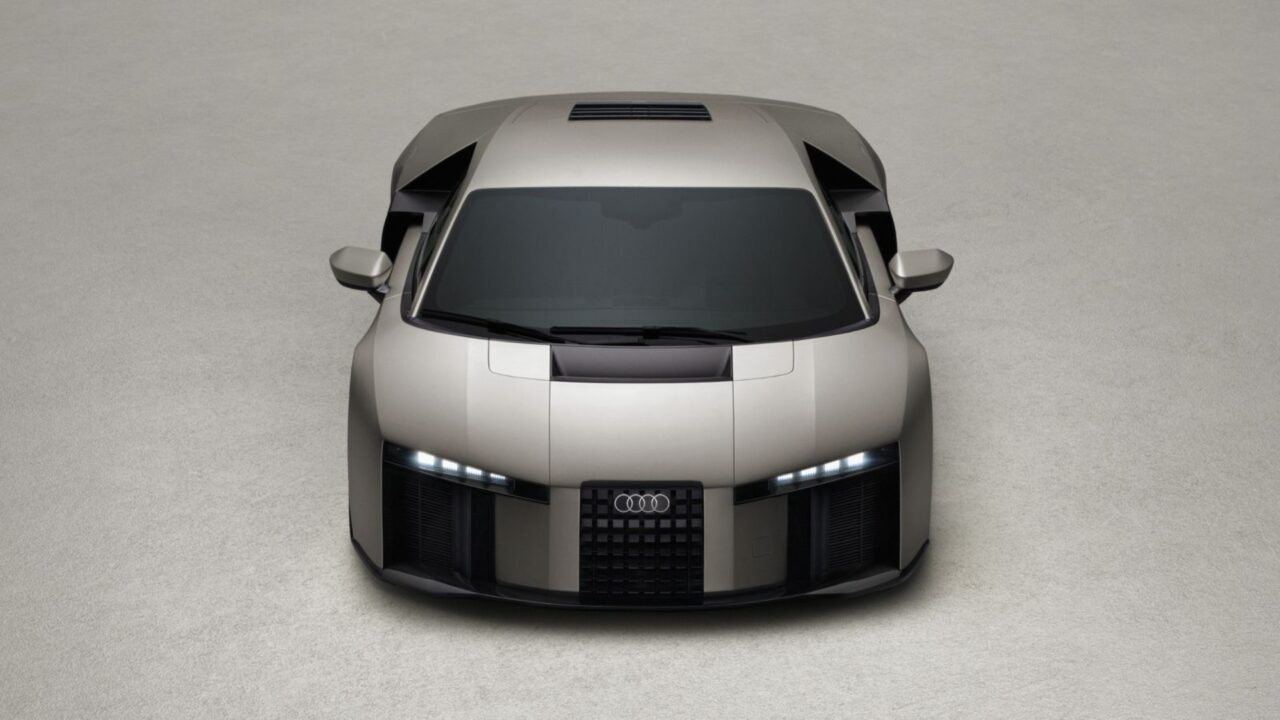 Vista frontal superior do Audi Nuvolari Concept em tom chumbo, fotografado em estúdio com fundo cinza neutro. O veículo apresenta carroceria de linhas esportivas e angulares, para-brisa amplo e escurecido, teto com abertura solar