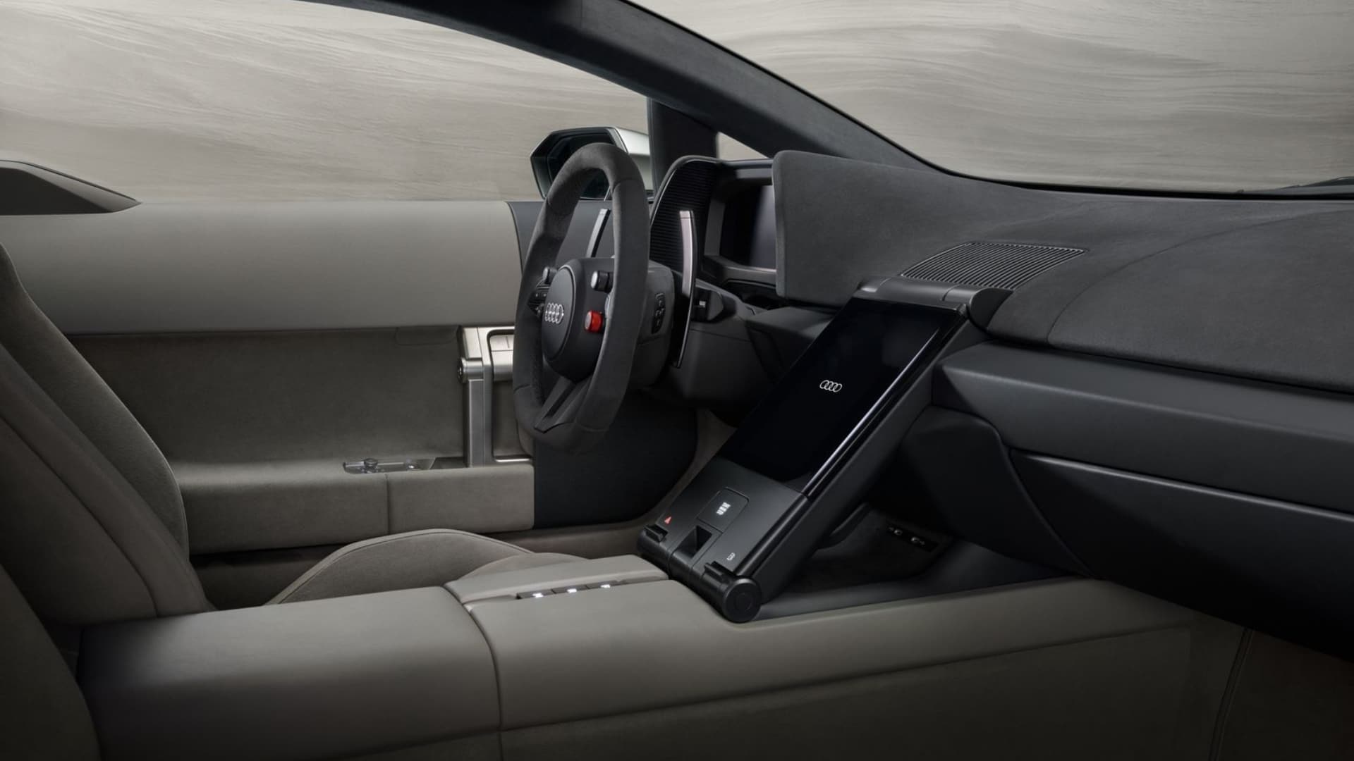 Texto alternativo: Interior do Audi Nuvolari Concept com acabamentos em couro e tecido cinza-escuro. Destaque para o volante esportivo com logotipo Audi e botão vermelho, painel de instrumentos digital e tela central inclinada com os quatro anéis da marca