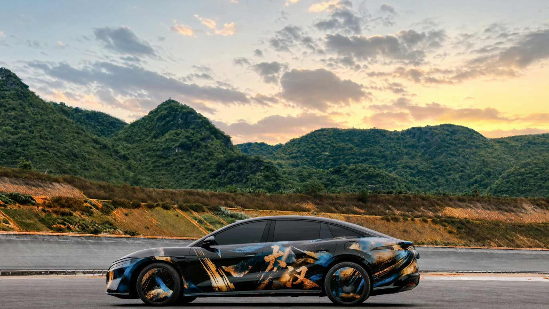 Vista lateral do BYD Grand Han com envelopamento artístico em preto, azul e dourado com caligrafia chinesa, estacionado em pista ao pôr do sol com montanha ao fundo