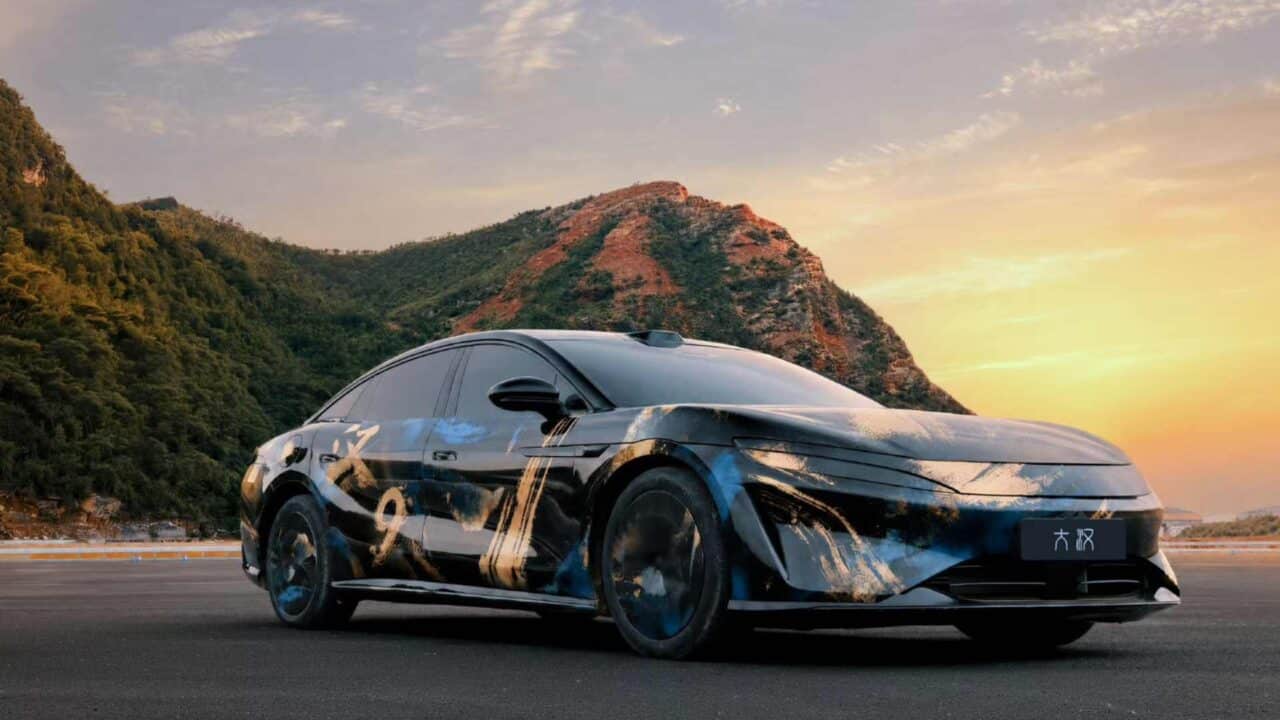 Vista frontal em ângulo três quartos do BYD Grand Han com envelopamento artístico em preto, azul e dourado com caligrafia chinesa, estacionado em pista ao pôr do sol com montanha ao fundo