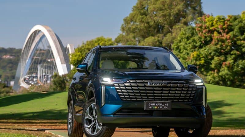 SUV GWM Haval H6 híbrido flex azul escuro estacionado em área verde, com ponte em arco ao fundo e árvores ao redor