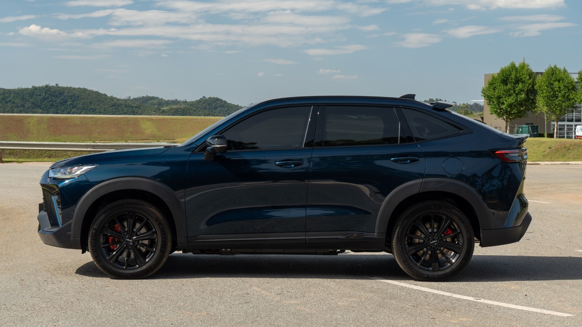 Vista lateral do GWM Haval H6 híbrido flex 2027 na cor azul escuro, com rodas pretas de liga leve, pinças de freio vermelhas, teto com spoiler integrado e vidros escurecidos, estacionado em área aberta com vegetação ao fundo