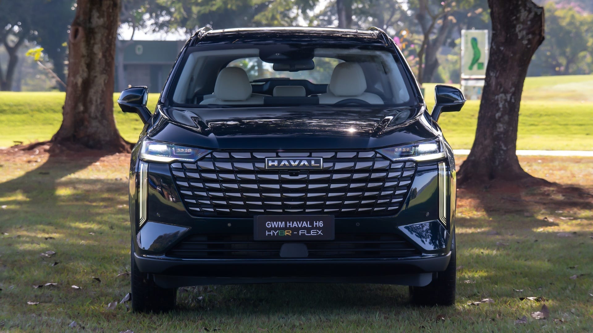 Vista frontal do GWM Haval H6 Hybr-Flex 2027 na cor preta, destacando a grade dianteira de design xadrez com logo Haval centralizado, faróis LED e placa identificando o modelo híbrido flex, estacionado em área gramada arborizada