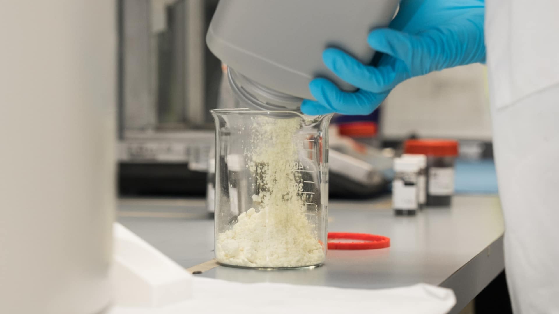 Mão com luva azul despeja enxofre em pó de cor clara dentro de um béquer de vidro em um laboratório. Ao fundo, equipamentos e frascos desfocados compõem o ambiente de pesquisa científica