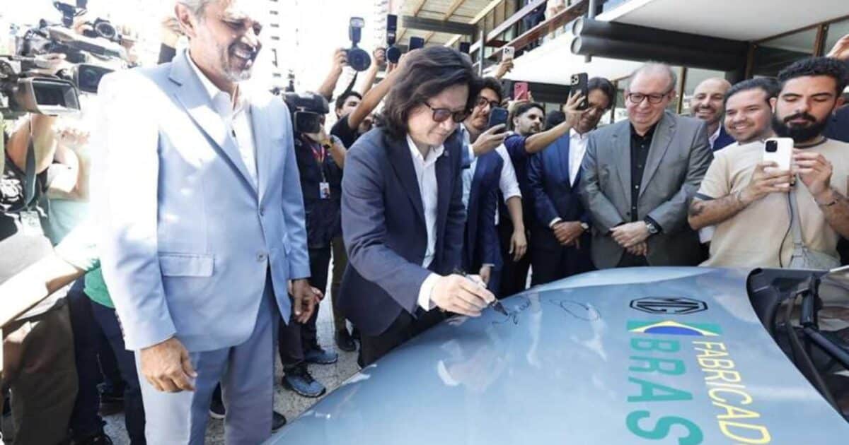 Executivos da MG Motor assinam o capô de um carro durante evento que anunciou a produção da marca no Ceará, cercados por autoridades, convidados e fotógrafos