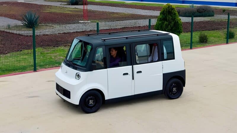 Veículo elétrico compacto Olinia 1, na cor branca com teto preto, estacionado em área pavimentada, com formato quadrado e capacidade para até seis ocupantes