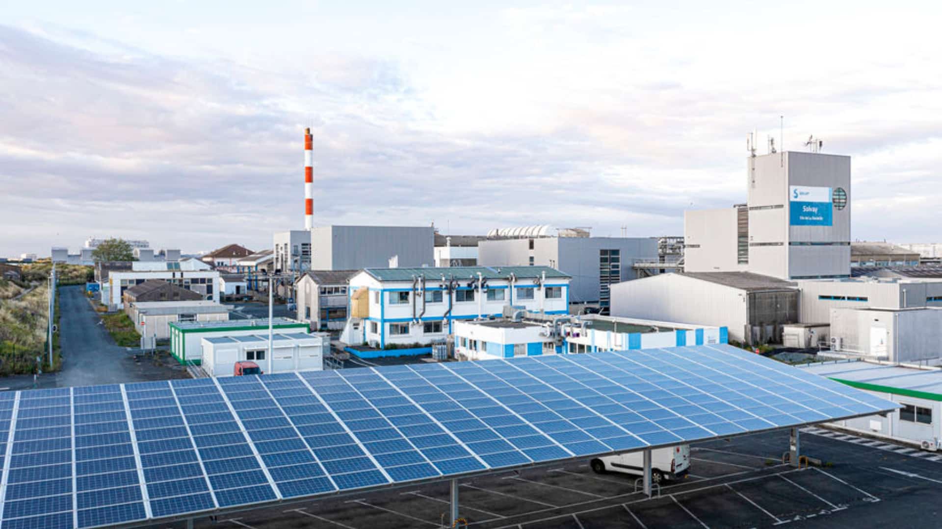 Fábrica da Solvay em La Rochelle, França, com painéis solares no primeiro plano e chaminé industrial ao fundo