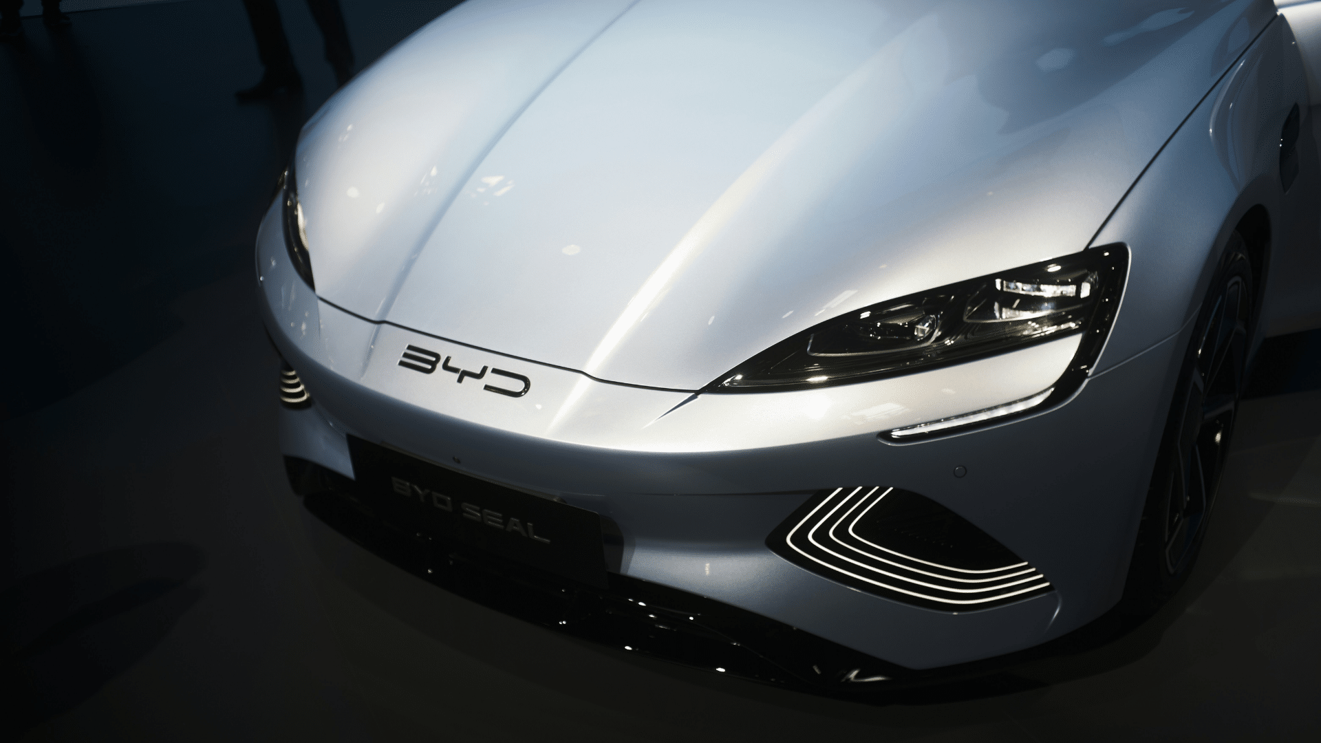 Detalhe da dianteira do carro elétrico branco BYD Seal, destacando seus faróis e logotipo em um showroom escuro