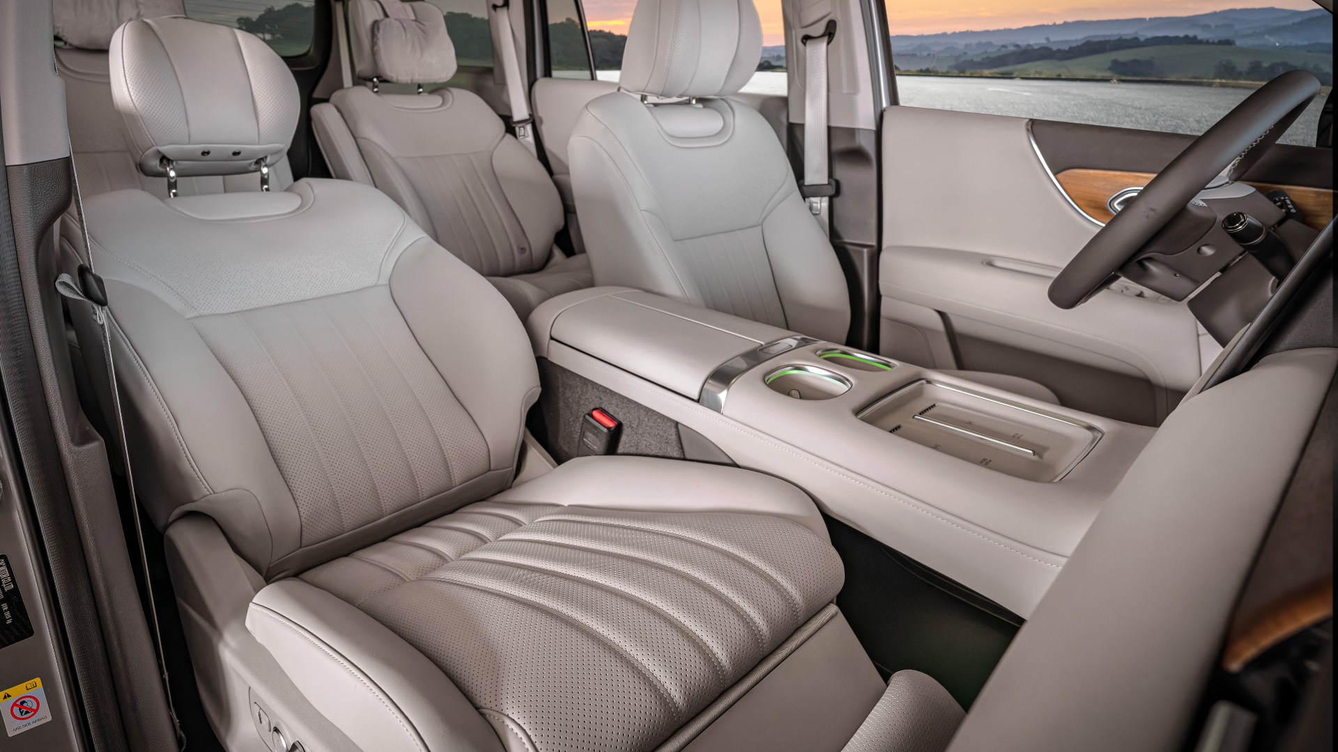 Interior luxuoso do GAC GS9, com foco no assento do passageiro revestido em couro perfurado de tom claro. O console central amplo, os acabamentos em madeira nobre e a iluminação ambiente verde complementam o design premium, com vista para um cenário de pôr do sol pela janela