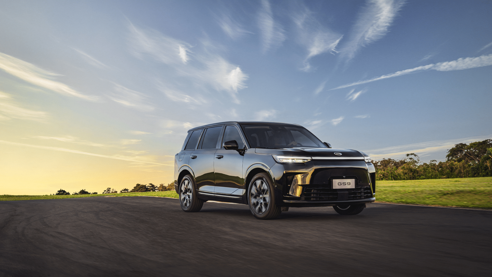 SUV preto de grande porte, modelo GAC GS9, em movimento sobre uma estrada de asfalto. Ao fundo, um pôr do sol com nuvens alongadas no céu azul e um campo verdejante