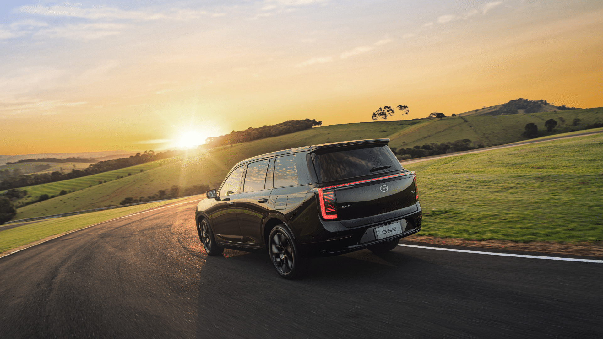 Traseira do SUV preto GAC GS9 trafegando em uma pista de asfalto. A foto é tirada durante o pôr do sol, com a luz dourada incidindo diretamente sobre a estrada e as colinas ao fundo, enquanto a lanterna traseira em LED brilha intensamente.