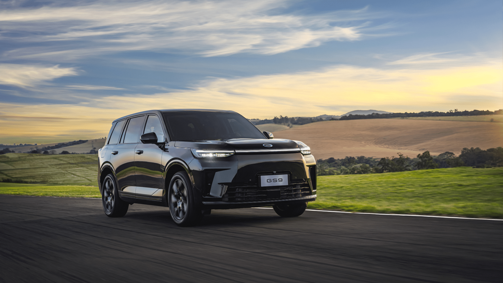 SUV preto GAC GS9 em movimento sobre uma estrada de asfalto. O veículo possui faróis dianteiros em LED e uma grande grade frontal. O cenário é composto por campos verdejantes, campos de cultivo e um céu com nuvens suaves no final da tarde
