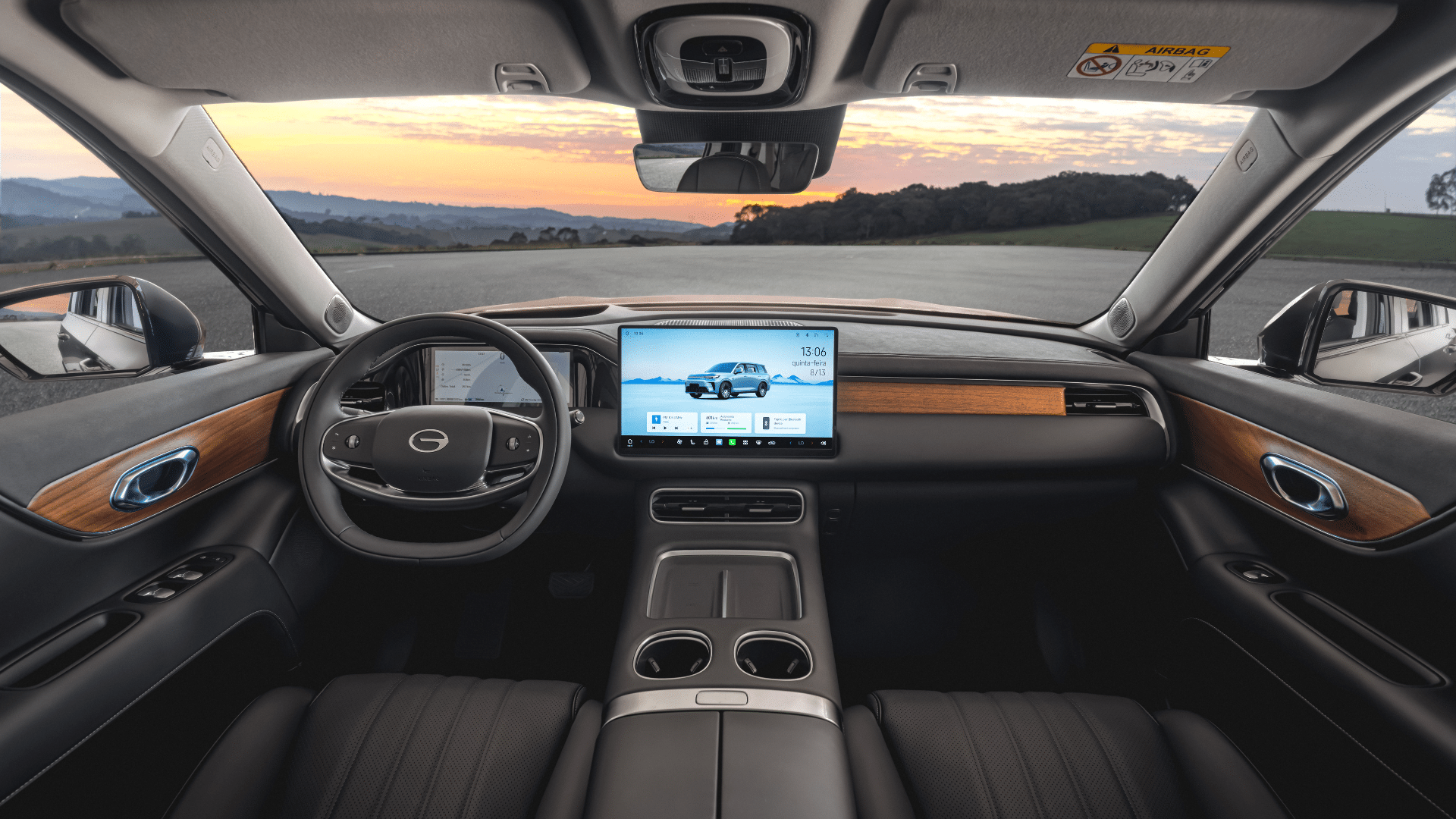 Interior do SUV com acabamento em preto, madeira nobre e detalhes cromados. O painel central exibe uma grande tela touch mostrando uma interface de clima e uma ilustração do carro. O volante escuro e o console central com porta-copos e carregador de celular são visíveis