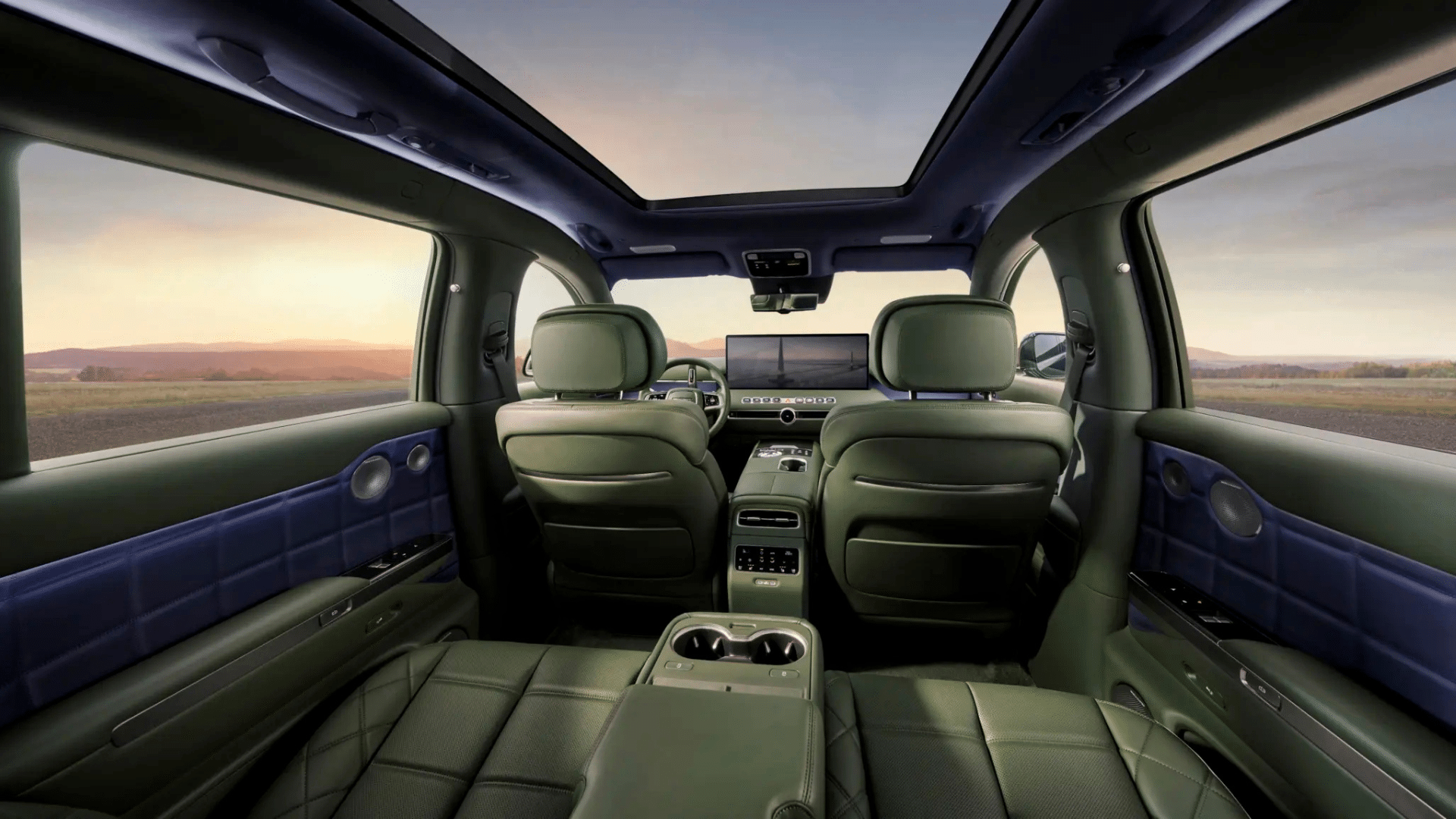 Vista dos bancos traseiros do Genesis GV90 revestidos em couro verde escuro, com teto solar panorâmico e laterais em painéis azuis. O amplo console central entre os assentos conta com porta-copos e painel de controle touchscreen. A vista externa revela um pôr do sol sobre montanhas