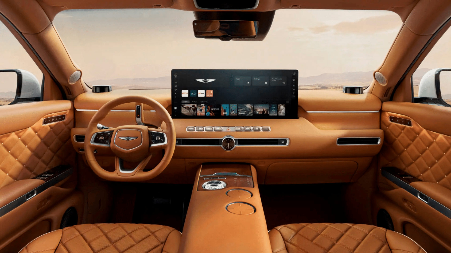 Interior do Genesis GV90 em couro caramelo com detalhes em pesponto losango. Destaque para o volante de dois raios com o logotipo da marca, uma ampla tela panorâmica exibindo aplicativos de mídia, saídas de ar horizontais e um console central alto com mostrador de relógio e controles touchscreen. Paisagem desértica visível pelo para-brisa