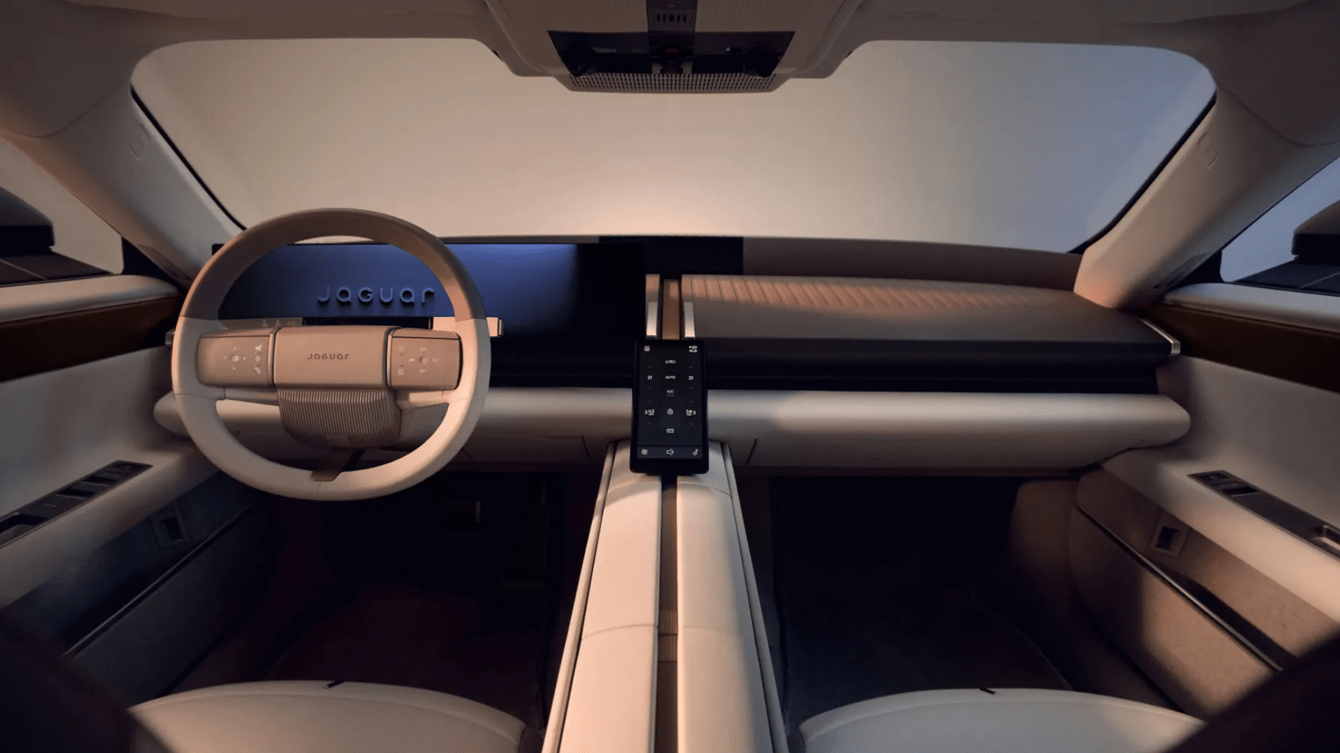 Vista do painel de instrumentos e volante do Jaguar Type 01. O volante é revestido em material claro, com o logotipo "JAGUAR" em destaque no centro e botões de toque integrados. À frente, uma grande tela digital escura exibe o nome da marca. O painel é dividido por uma linha central vertical que desce pelo console, onde se encontra uma tela central de controle, também vertical, com ícones iluminados. Acabamentos em madeira escura e tecido claro compõem o visual minimalista