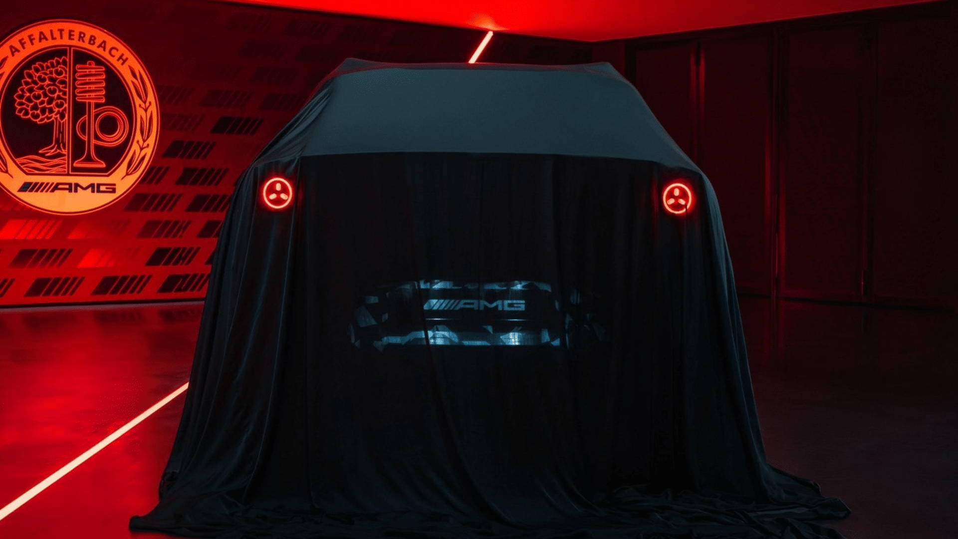Parte traseira do SUV elétrico da Mercedes-AMG sob uma capa preta. As lanternas traseiras em formato circular emitem uma luz vermelha intensa. No centro da traseira, o logotipo "AMG" brilha em azul e branco sobre o tecido, em um ambiente com fortes reflexos vermelhos no chão.