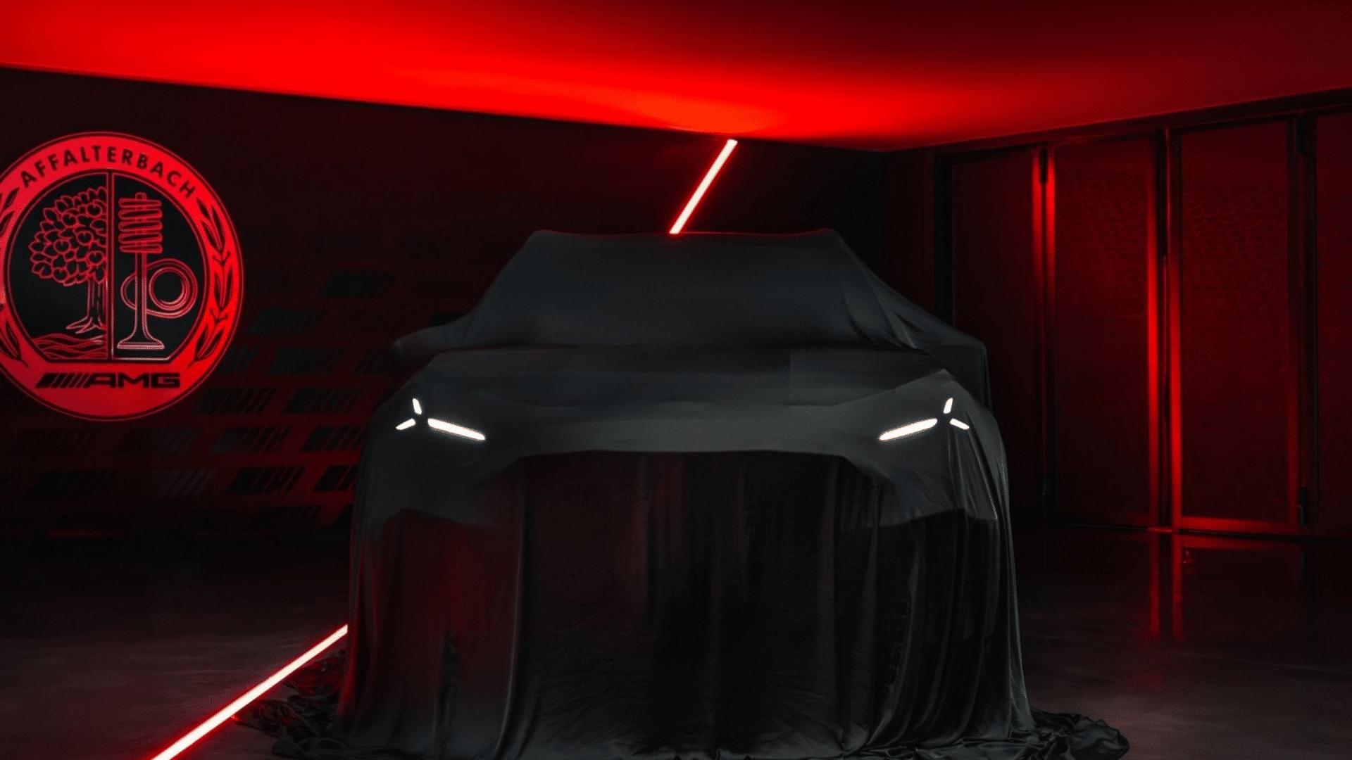 Carro coberto por um pano preto em um ambiente escuro com iluminação vermelha dramática. Apenas os faróis em formato de fenda iluminados em branco são visíveis, destacando a silhueta do SUV. Ao fundo, o logotipo circular vermelho da Mercedes-AMG com a inscrição "Affalterbach".