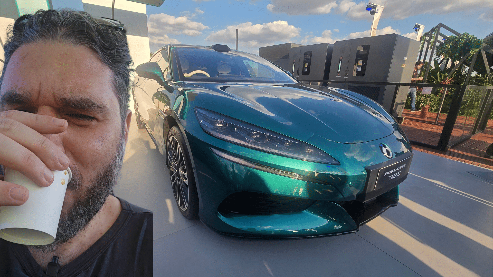 O jornalista Fábio Marton segurando um copo de café em frente ao Denza Z9 GT e o carregador BYD Flash