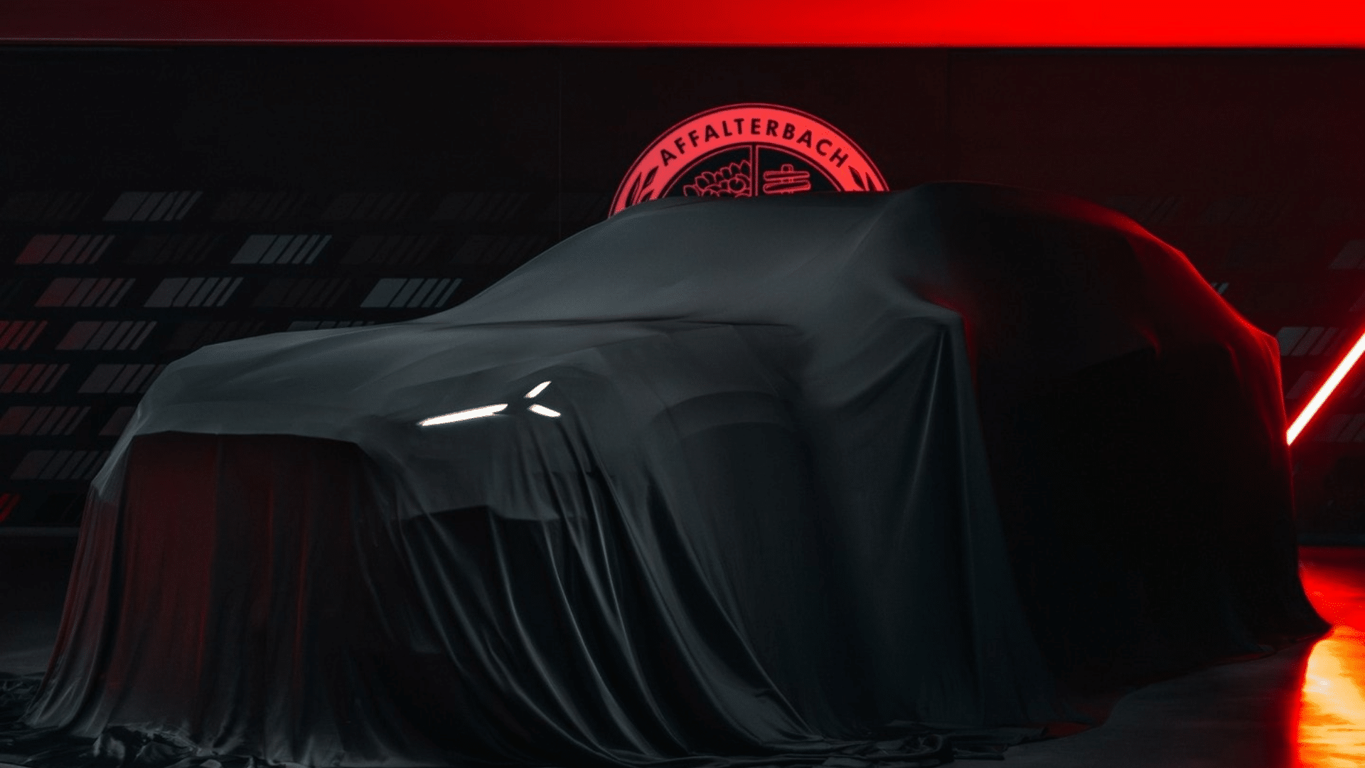 Vista frontal de um SUV da Mercedes-AMG totalmente encoberto por um tecido preto. A iluminação vermelha do estúdio contrasta com os faróis dianteiros em LED branco. À esquerda, na parede, um grande emblema luminoso vermelho da AMG com o símbolo da árvore e do pistão.