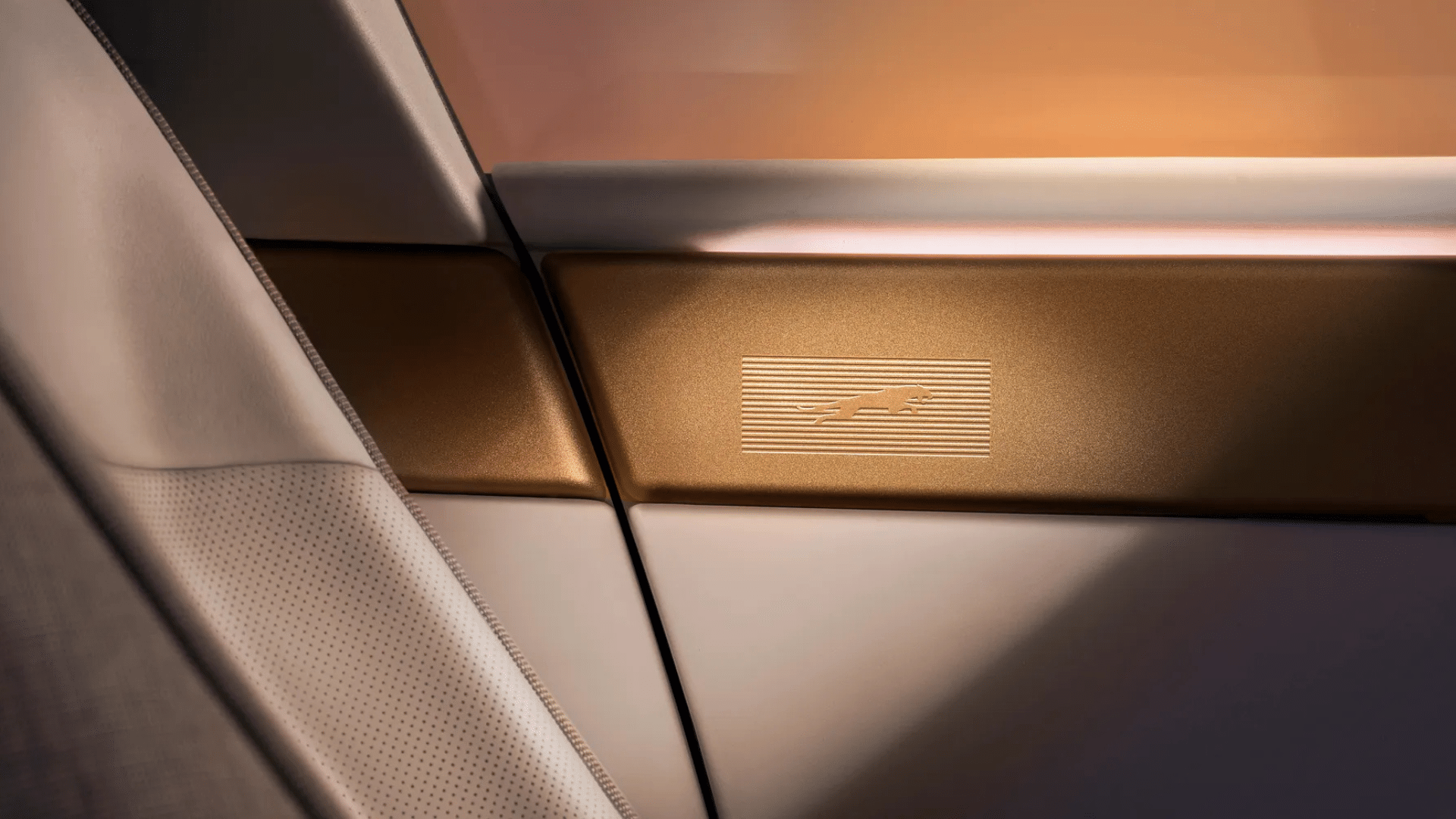 Detalhe de acabamento interno do Jaguar Type 01. Em uma superfície de metal com textura escovada e tom cobre-dourado, há um logotipo da Jaguar (a figura do jaguar saltando) gravado em baixo relevo, sobreposto a finas linhas horizontais. A peça metálica é margeada por um painel de couro ou tecido claro com perfurações precisas, destacando o requinte dos materiais.