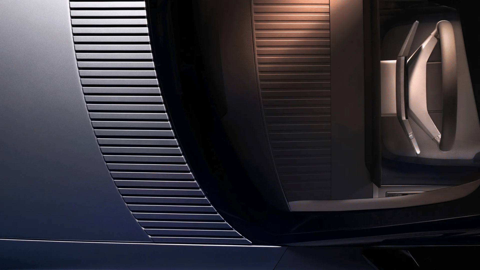 Close-up do interior do Jaguar Type 01 mostrando o painel da porta. Destaca-se um design geométrico com ripas horizontais em metal escuro que se curvam suavemente em direção ao acabamento inferior. Ao fundo, vê-se o encosto de um banco com uma alça de tecido minimalista, tudo sob uma iluminação suave que alterna entre tons frios de cinza e quentes de cobre.
