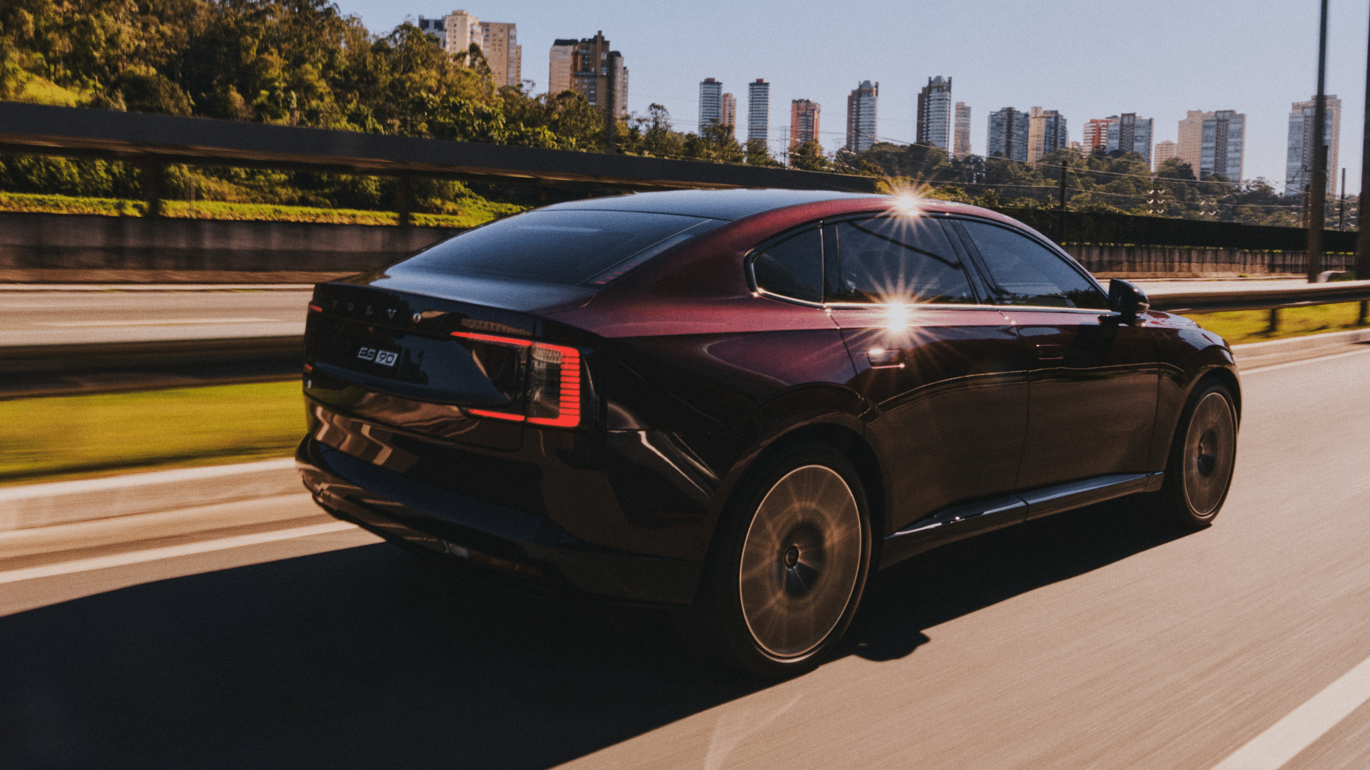 Vista traseira do Volvo ES90 em movimento na estrada, com desfoque de velocidade no fundo. A imagem evidencia o design fastback do sedã, as lanternas traseiras em LED com formato de 'C' alongado, a traseira limpa e minimalista, e o nome do modelo escrito no porta-malas sob o logotipo Volvo, refletindo a luz do entardecer.