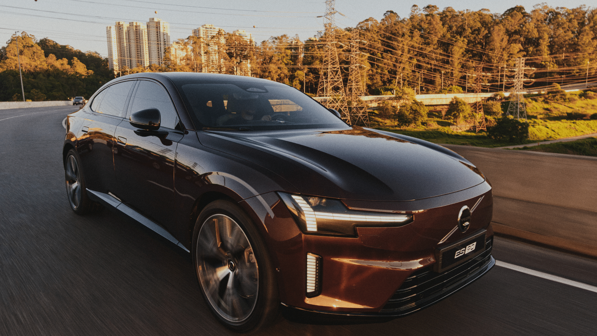 Volvo ES90 em movimento em uma rodovia durante o pôr do sol. A imagem mostra a vista lateral e frontal do sedã em tom vinho, destacando a fluidez do design escandinavo, as linhas aerodinâmicas da carroceria e a imponência da dianteira com luzes diurnas em LED acesas
