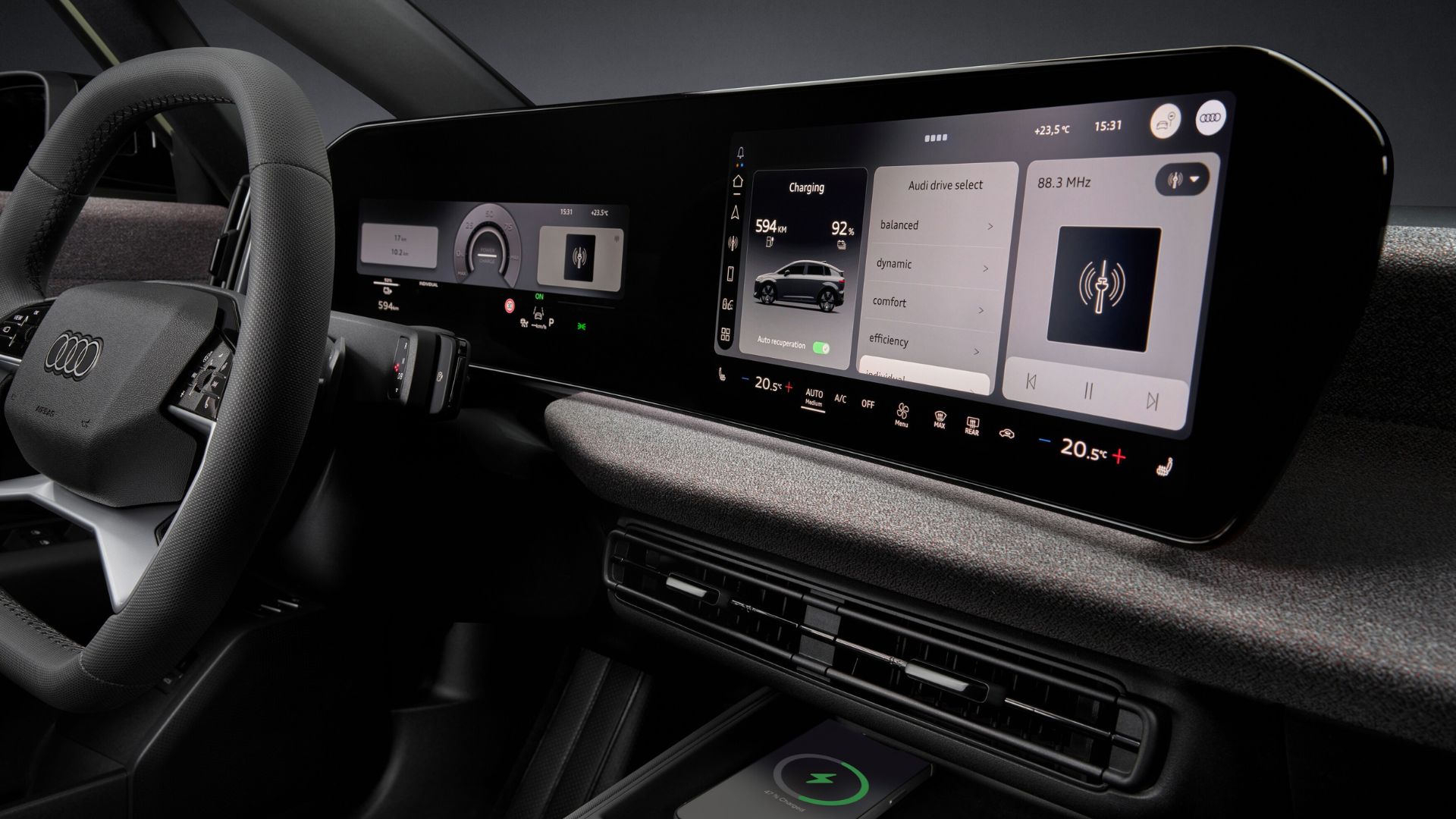 Close-up da tela dupla (instrumentos e multimídia) do Audi ligada. A tela exibe o aplicativo de carregamento (Charging) e o menu de modos de condução (Audi drive select). Abaixo da tela, encontra-se o console central com saídas de ar e uma base de carregamento sem fio para celular.