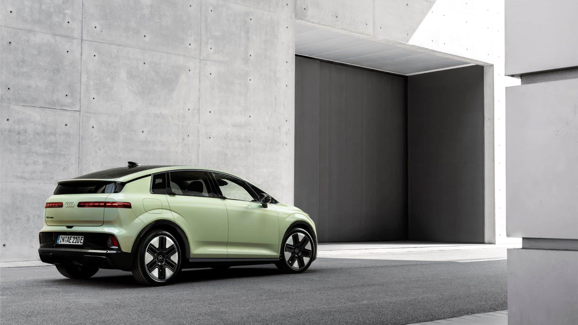 Vista traseira em ângulo de três quartos do Audi Q6 e-tron pintado em verde-claro (tom pastel). O carro está estacionado em um ambiente moderno de concreto, destacando a lanterna traseira em faixa de LED contínua e as rodas de liga leve com design aerodinâmico