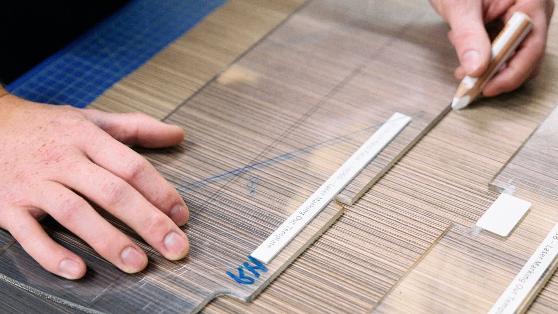 Mãos de um artesão trabalhando sobre uma bancada com uma folha de material compósito de fibra de carbono com padrão trançado, coberta por uma película protetora transparente e uma máscara azul