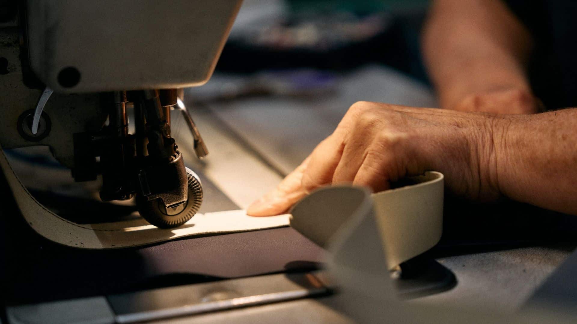 Close-up de uma máquina de costura industrial em operação, com a agulha descendo sobre uma tira de couro bege.