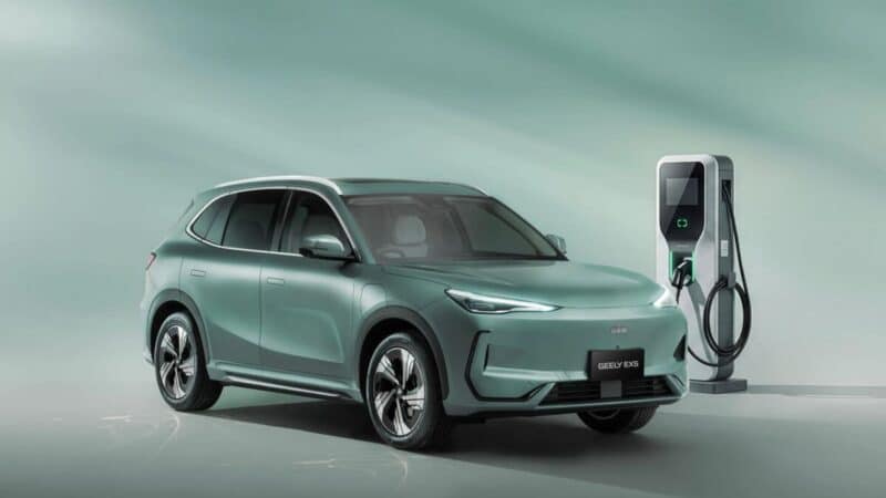 SUV elétrico da Geely verde-claro em um estúdio, estacionado ao lado de uma estação de carregamento elétrico