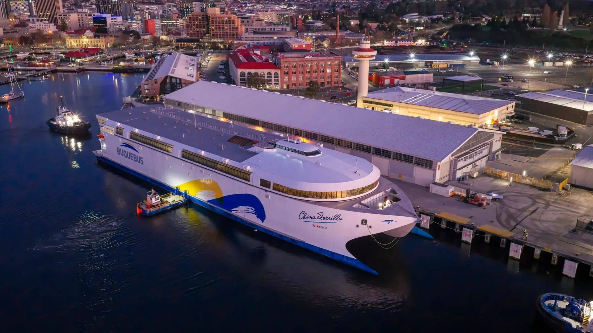Navio de passageiros China Zorrilla atracado no porto à noite. A foto aérea mostra o casco branco e azul do navio, o cais iluminado e os prédios históricos da cidade ao fundo