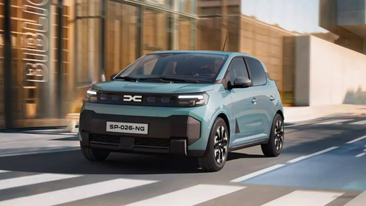 Dacia Spring verde-água em movimento pela cidade, capturado em ângulo frontal de três quartos. A imagem mostra levemente desfocada ao fundo para transmitir a sensação de velocidade. Destacam-se os vincos do capô, o farol dianteiro em LED e os detalhes em relevo no para-choque preto, além do contraste entre o carro e o asfalto com faixas de pedestres