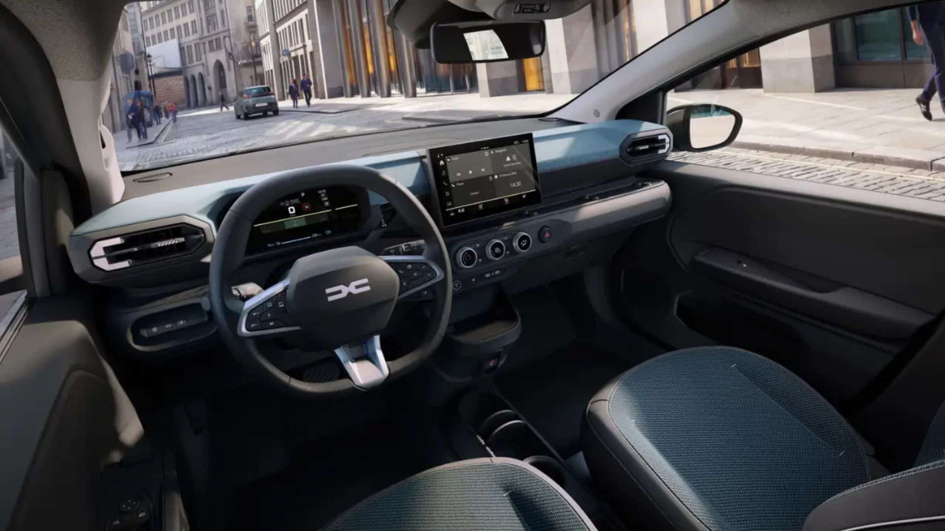 Interior do Dacia Spring visto do banco do motorista. O painel é escuro com um acabamento horizontal em tom azul-acinzentado na parte superior. Possui volante de três raios com o logo central, painel de instrumentos digital atrás do volante e uma tela multimídia flutuante no centro