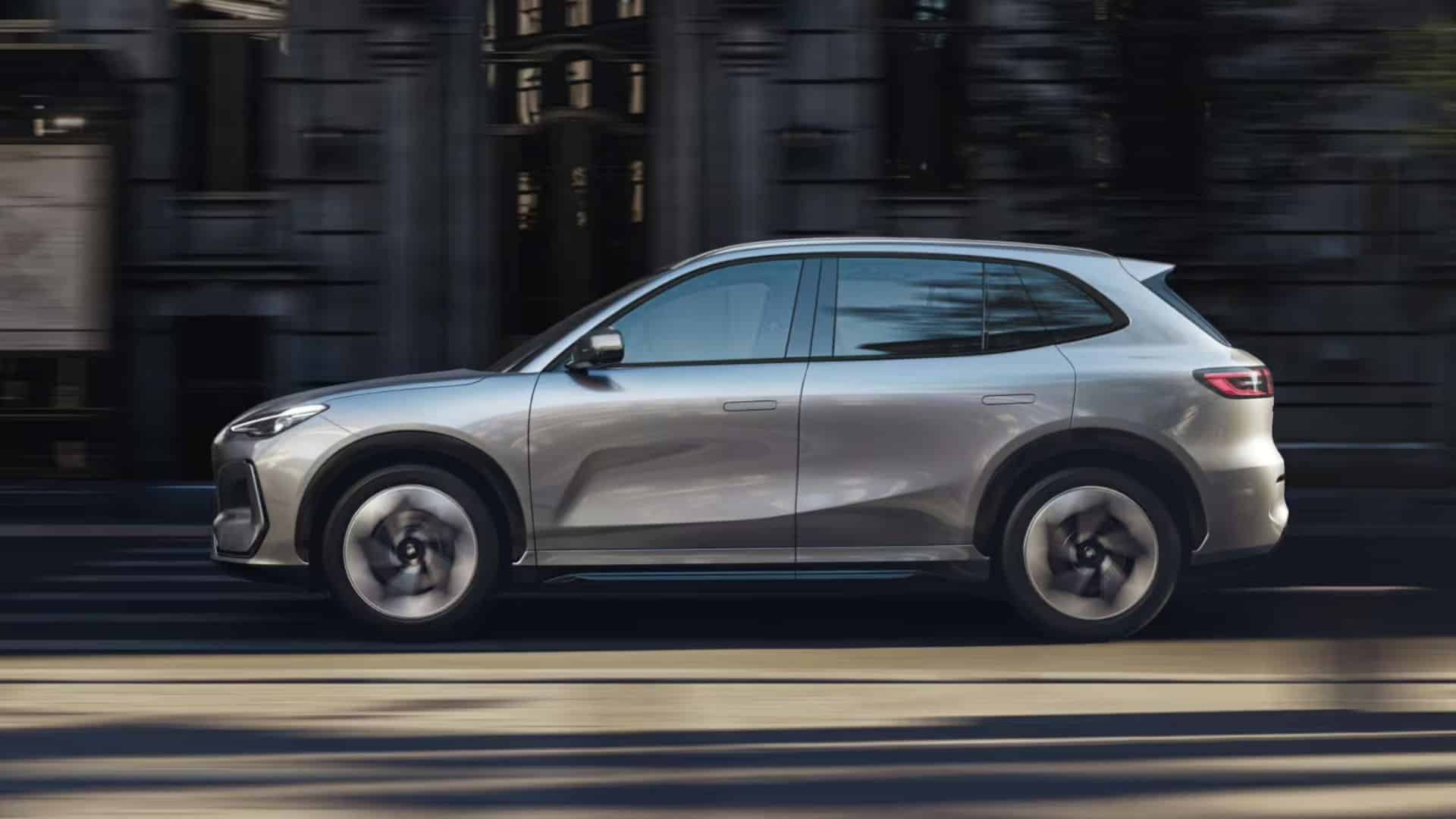 Vista lateral do Geely Galaxy E5 prateado em movimento por uma rua urbana, com desfoque de movimento nas rodas e no fundo de prédios escuros, destacando o design aerodinâmico e as rodas com detalhes prateados