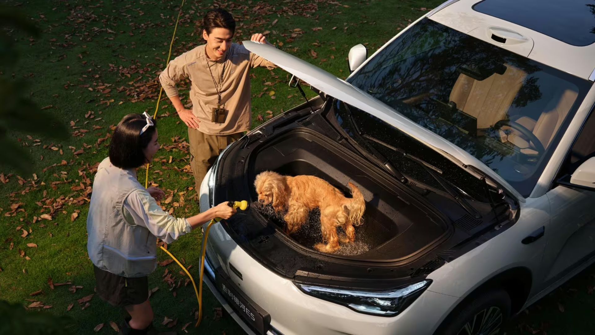 Vista superior do Geely Galaxy E5 branco com o capô aberto, revelando um cachorro dentro do compartimento dianteiro (frunk) sendo lavado com uma mangueira por uma mulher, enquanto um homem observa. O carro está estacionado em um gramado com folhas secas
