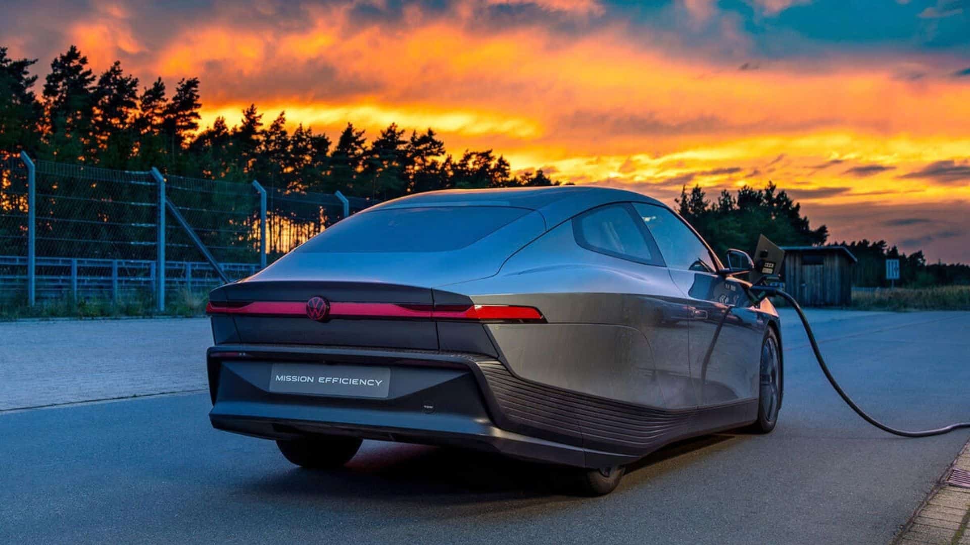 Volkswagen Mission Efficiency estacionado e conectado a uma estação de carregamento. A cena ocorre ao anoitecer, com um céu alaranjado vibrante ao fundo, destacando a traseira do veículo elétrico e o cabo de carregamento conectado à lateral