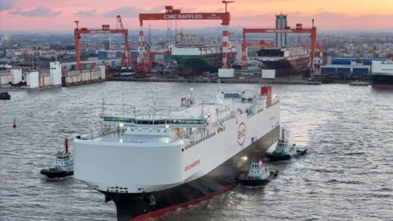 Fotografia de um enorme navio de carga branco com a inscrição BYD na lateral, sendo manobrado por pequenos rebocadores em um canal portuário movimentado, com o céu tingido de rosa e laranja