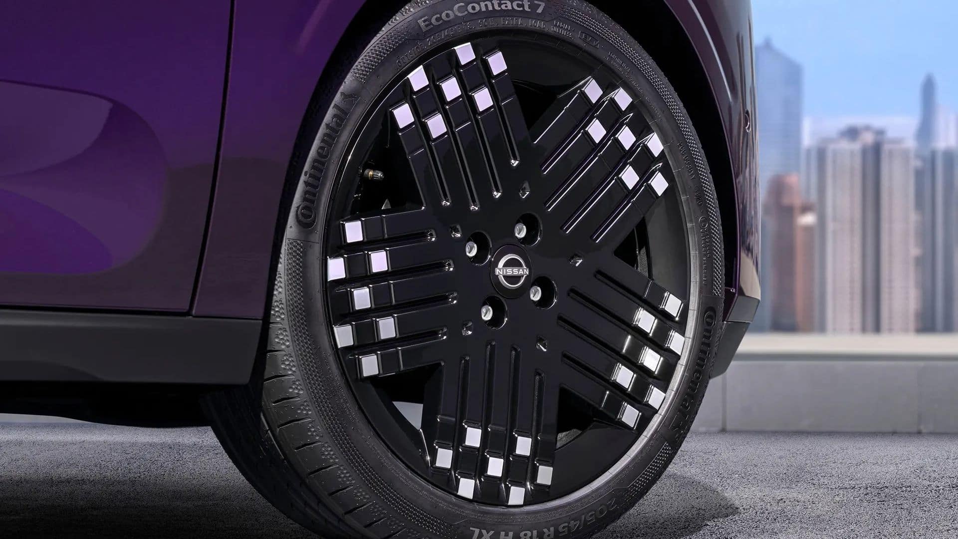Detalhe da roda dianteira do Nissan PIXO, com calota preta brilhante apresentando detalhes quadrados prateados e o logotipo Nissan ao centro. O pneu é um Continental EcoContact 7, com a inscrição '205/45 R18' visível na lateral