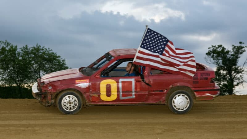 Carro muito antigo em prova de demolition derby com piloto levando bandeira dos EUA