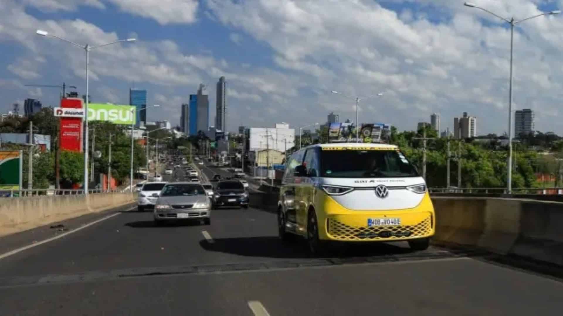 VW ID. Buzz amarelo e branco trafegando em uma rodovia movimentada de uma cidade não identificada, com arranha-céus ao fundo sob céu azul com nuvens