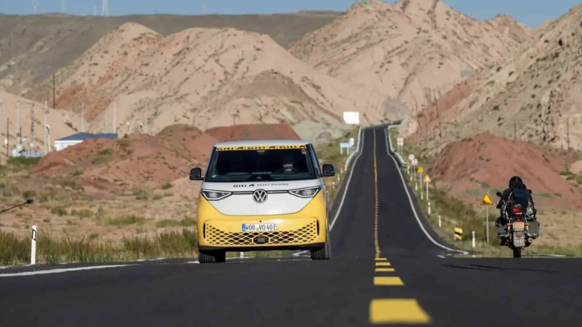 VW ID. Buzz na viagem recordista de Rainer Zietlow, na China. 
VW ID. Buzz amarelo e branco dirigindo em uma estrada asfaltada e sinuosa que corta uma paisagem árida de colinas avermelhadas e vegetação rasteira, sob céu clara